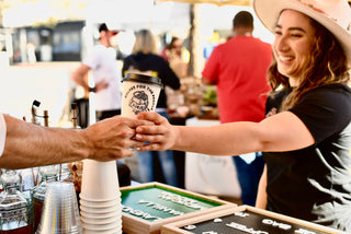 Photo was taken at the pop-up cafe in North Park, San Diego. It shows Surje Coffee (said like Surge Coffee) founder handing a cup of hot Ethiopia coffee. The coffee cup says "Coffee for the People | Surje Coffee Co" and shows a knapsack of coffee beans.